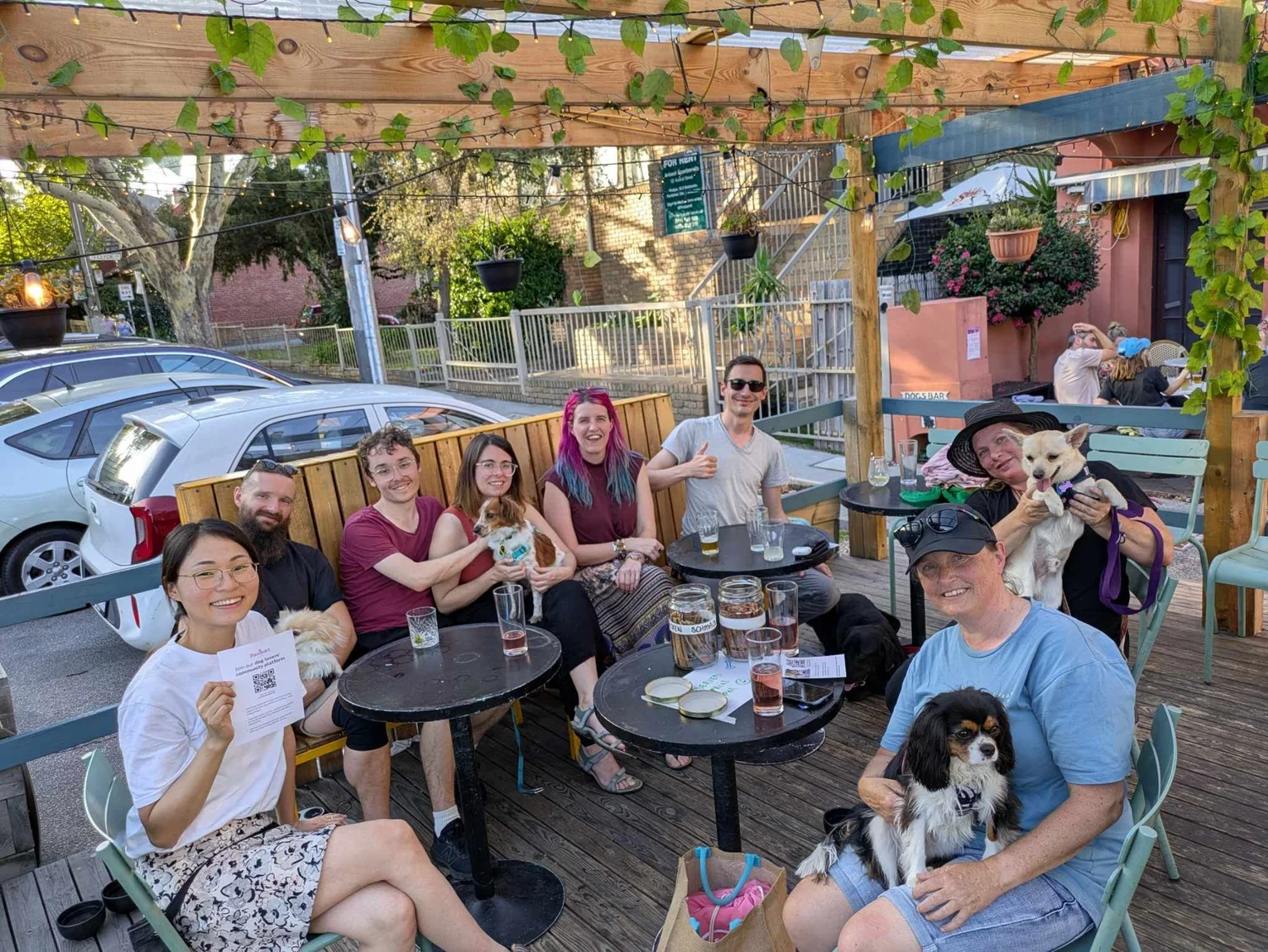 Pawshare Club members with dogs seated at outdoor cafe tables beneath hanging plants